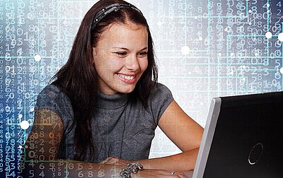 junge, sitzende, lächelnde Frau mit braunen langen Haaren sieht auf den Bildschirm des Laptops der vor ihr steht