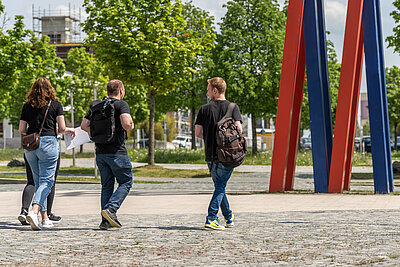 Foto vom Campus Hof, Studierende gehen an der Stele vorbei über den Alfons-Goppel-Platz