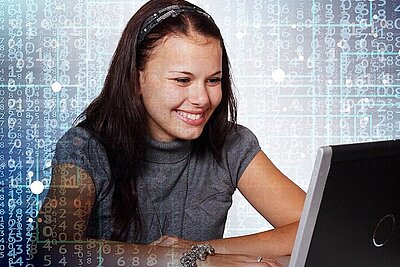 junge, sitzende, lächelnde Frau mit braunen langen Haaren sieht auf den Bildschirm des Laptops der vor ihr steht