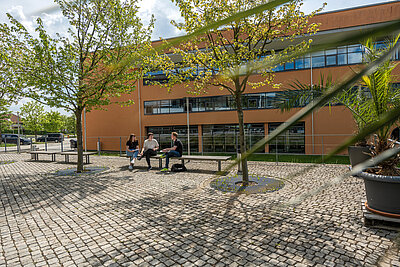 Blick auf das Gebäude A der Hochschule Hof; 3 Studierende sitzen auf einer Bank